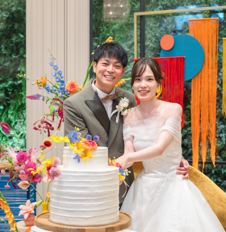 結婚式でウェディングケーキの前に立つ新郎新婦。ケーキは白色で、色とりどりの花が飾られている。