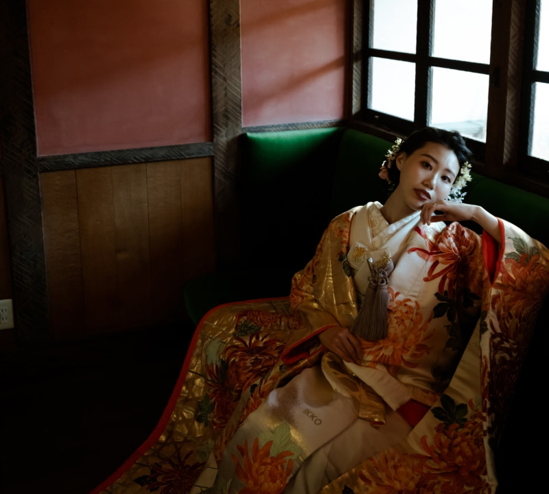 金色とクリーム色の豪華な着物を着た若い女性が、緑のソファに座ってカメラを見つめている。着物にはオレンジと赤の菊の模様が描かれ、髪には白い花の飾りが付いている