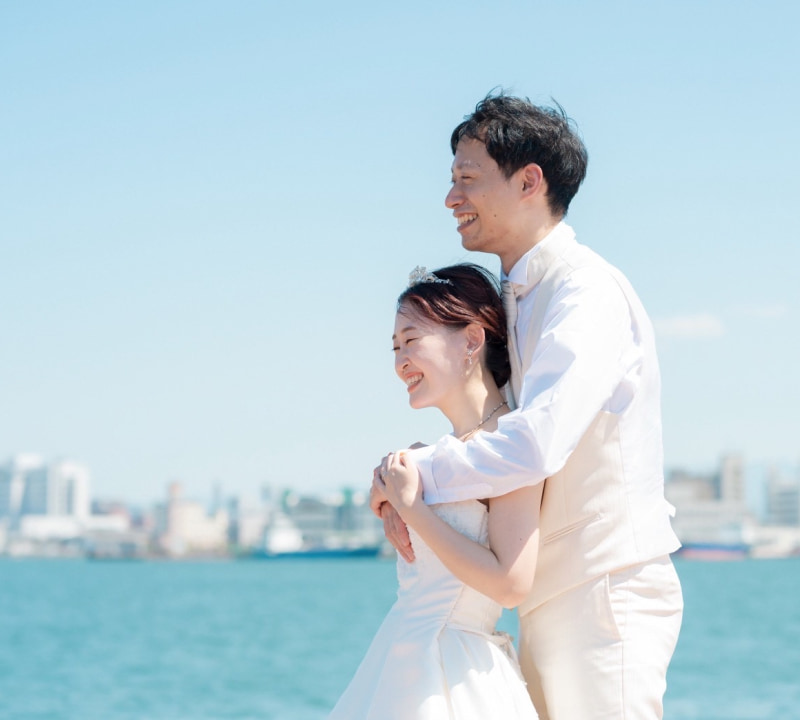 白いウェディングドレスを着た花嫁とクリーム色のタキシードを着た新郎が、青い海と都市のスカイラインを背景に、満面の笑みを浮かべて抱き合っている様子