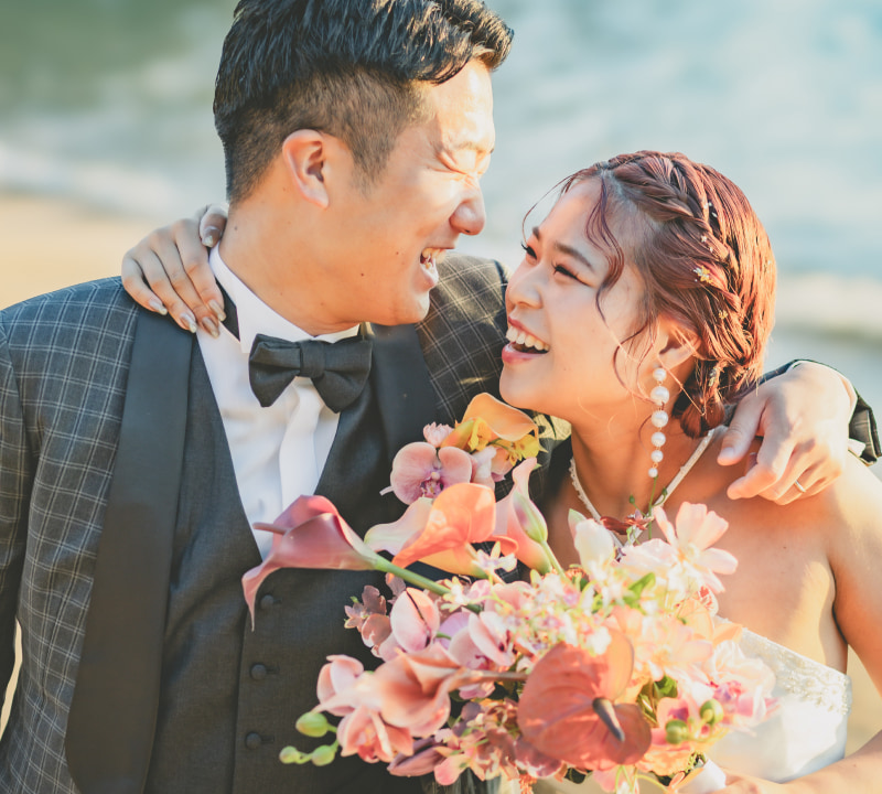 晴れた日のビーチで抱き合い、満面の笑みを浮かべている新郎新婦のクローズアップ。グレーのチェック柄スーツを着た新郎と白いストラップレスのウェディングドレスを着た花嫁が互いを見つめ合い、花嫁は鮮やかなピンク、ピーチ、オレンジ、深い赤の花々で構成されたブーケを手に持っている