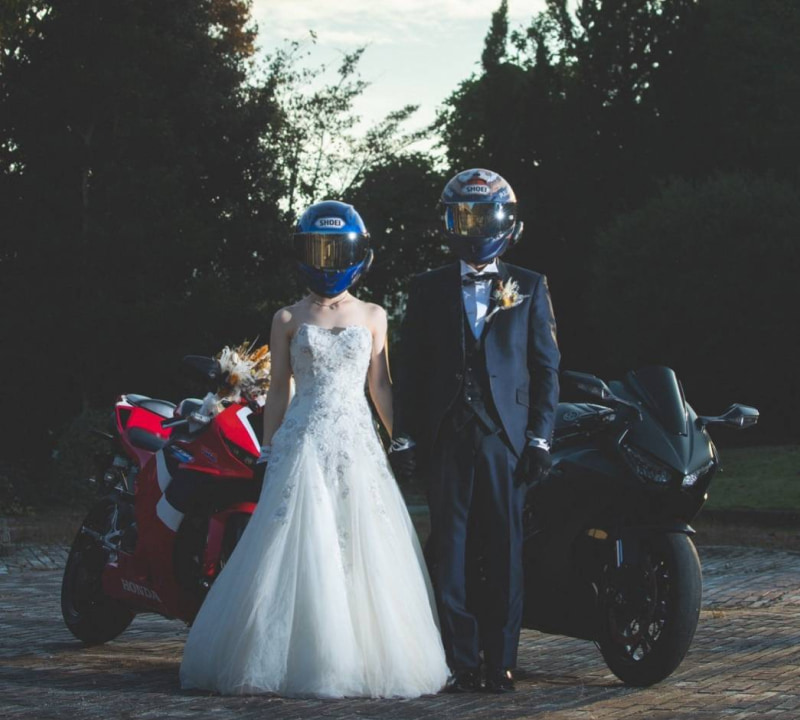 ウェディングドレスを着た新婦とタキシードを着た新郎がヘルメットをかぶり、バイクの横に立っているシーン。