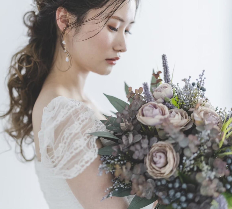 白いウェディングドレスを着た花嫁が、淡い紫とクリーム色の花々で構成されたブーケを優しく見つめているポートレート