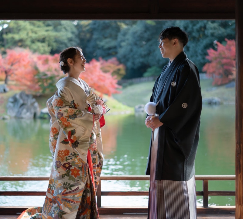 伝統的な和装の新郎新婦が、紅葉が美しい庭園の池を望む木造の回廊で、互いに見つめ合い笑顔を交わしている様子。花嫁はクリーム色の豪華な打掛を着用し、扇子を持っている
