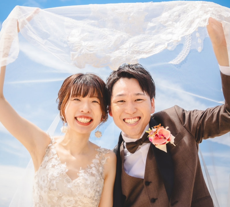 ウェディングドレスを着た新婦とタキシードを着た新郎が、青空の下でベールを持ち上げて笑顔を見せている