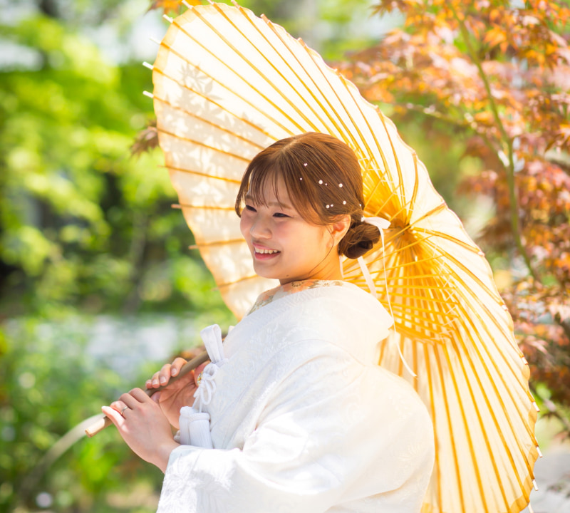 黄色い和傘を持つ女性が白い着物を着て微笑んでいる。背景には緑の植物が見える。
