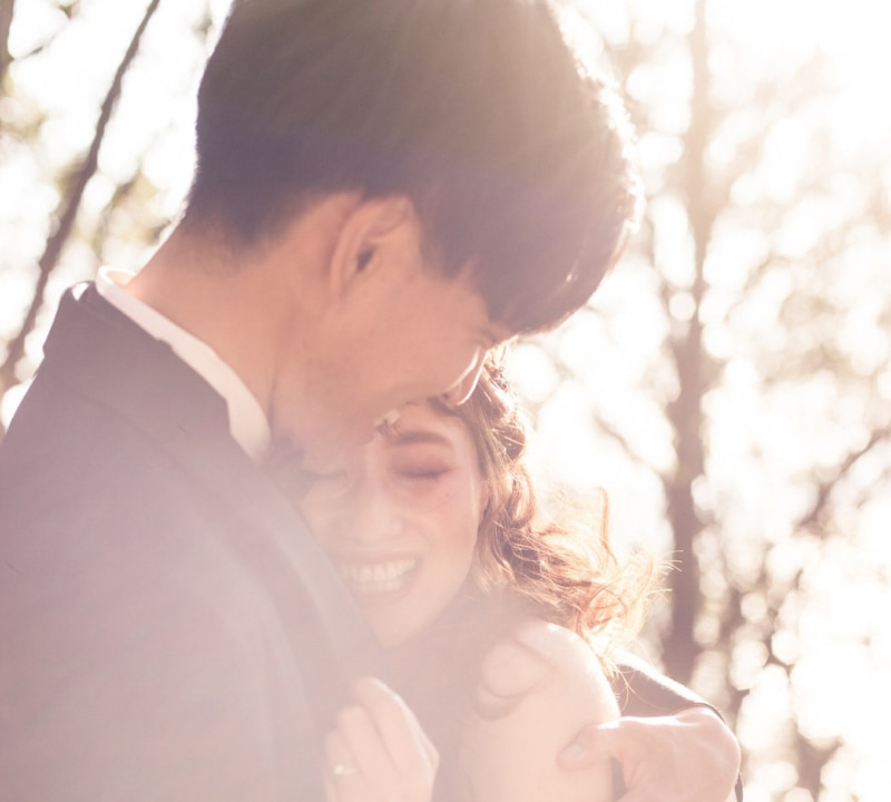 明るい光に包まれ、抱き合いながら互いを見つめ合う新郎新婦のクローズアップ。ダークなスーツを着た新郎と、オフショルダーの白いウェディングドレスを着た花嫁が満面の笑みを浮かべ、背景は柔らかくぼかされた屋外の風景が映えている