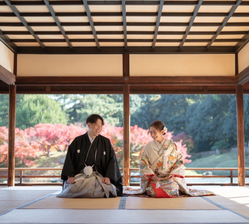 伝統的な日本の木造建築の畳敷きの屋内で、和装の新郎新婦が向き合って座り、笑顔を交わしている様子。背景には鮮やかな紅葉が広がる美しい日本庭園が見える