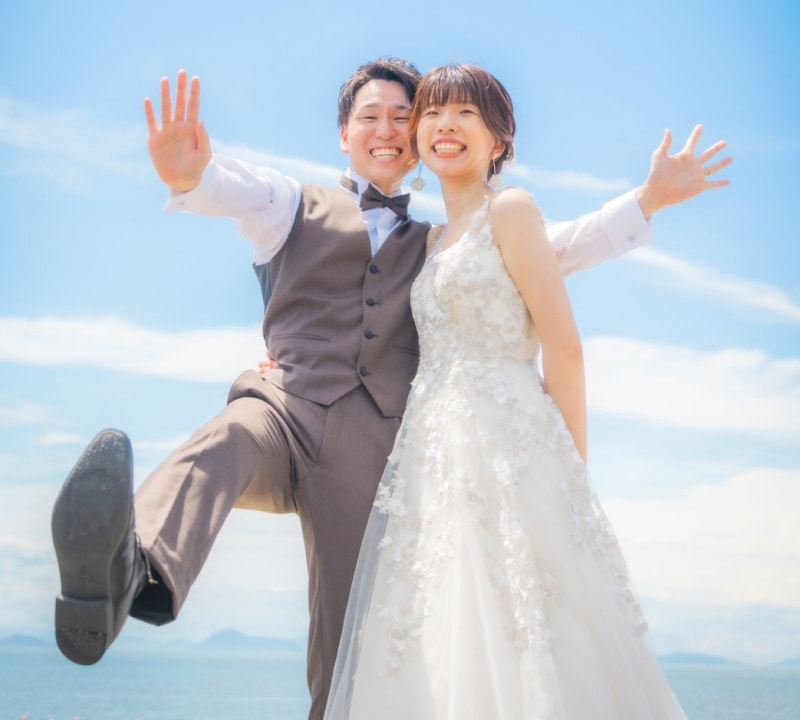 ウェディングドレスを着た新婦とタキシードを着た新郎が笑顔でポーズをとる。青空の下で楽しそうに立っている。