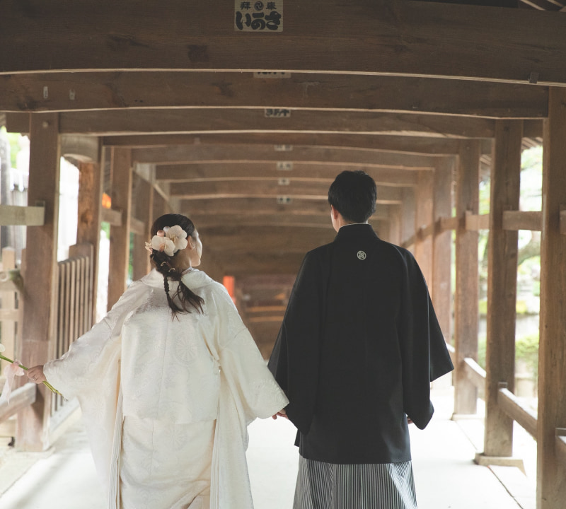 白無垢を着た女性と黒い着物を着た男性が手をつないでいる様子。伝統的な日本の結婚式の衣装を着た二人。
