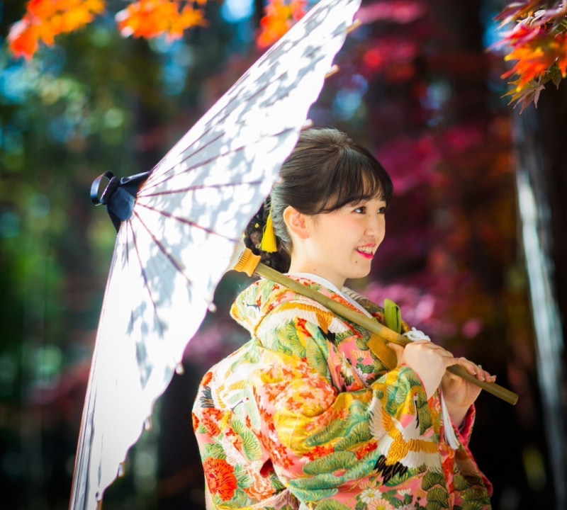 色とりどりの着物を着た女性が、和傘を持ちながら微笑んでいる。背景には紅葉が見える。