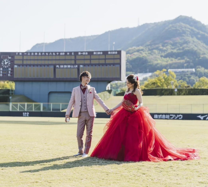 赤いウェディングドレスを着た花嫁とストライプのスーツを着た新郎が、野球場で手をつないで歩いている様子。花嫁は野球のグローブを持っている