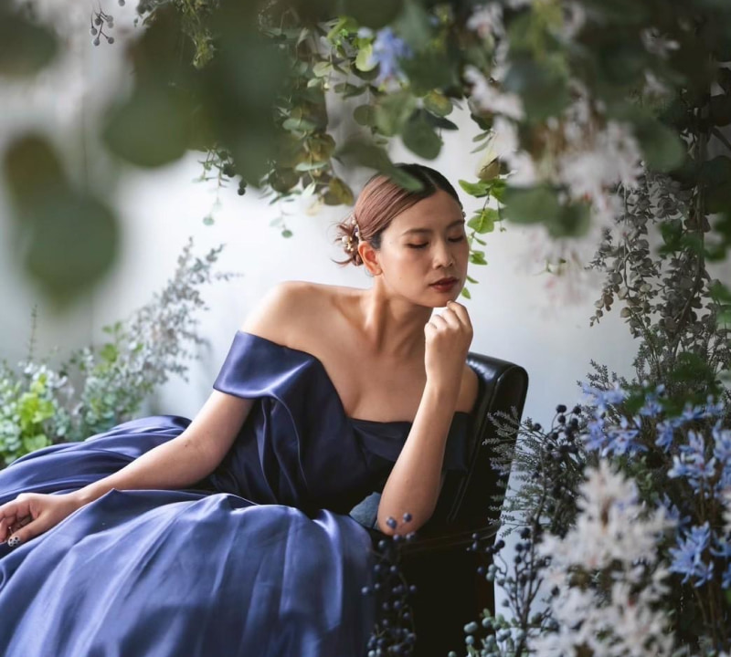 濃いネイビーブルーのオフショルダードレスを着た女性が、緑豊かな植物と花々に囲まれた中で椅子に座り、目を閉じて物思いにふけっている様子