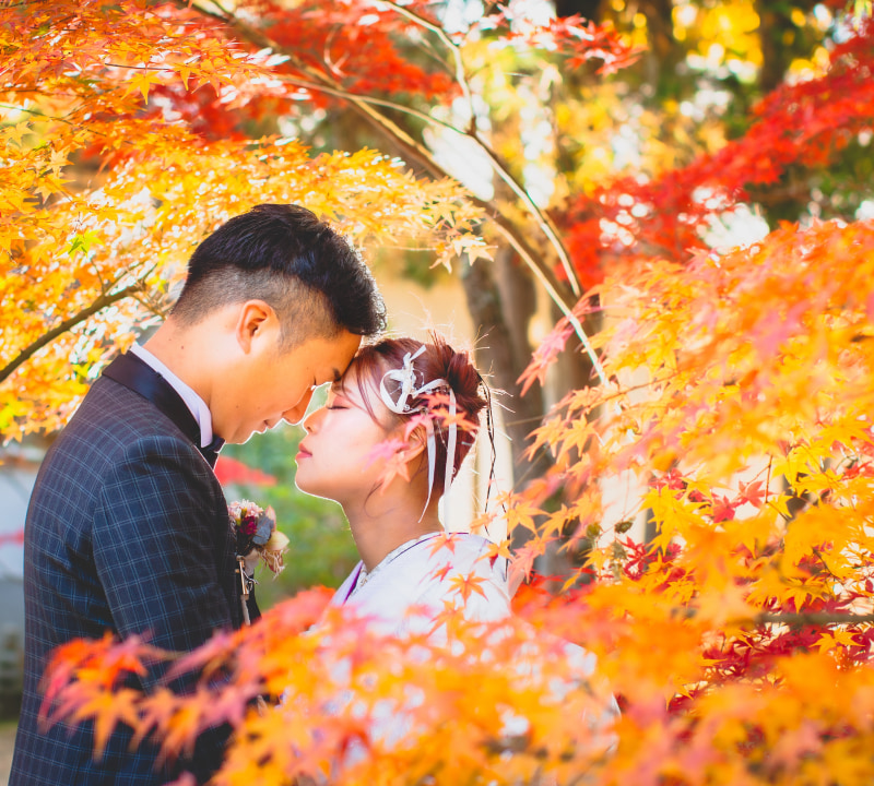 秋の紅葉の中で顔を寄せ合う新郎新婦。鮮やかな赤やオレンジの葉が背景に広がるシーン。
