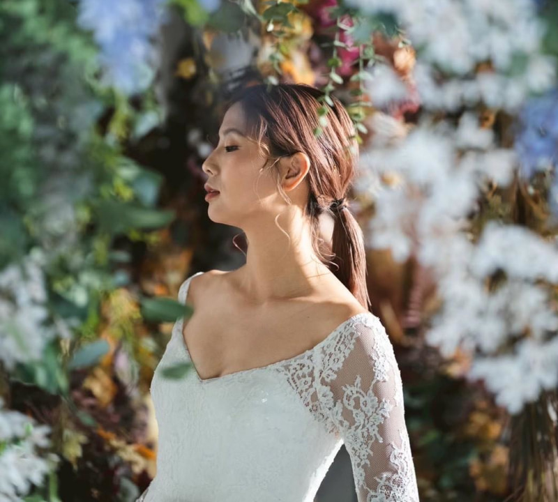 白いレースのウェディングドレスを着た花嫁が、花と緑に囲まれた中で横顔を見せ、穏やかな表情を浮かべているポートレート